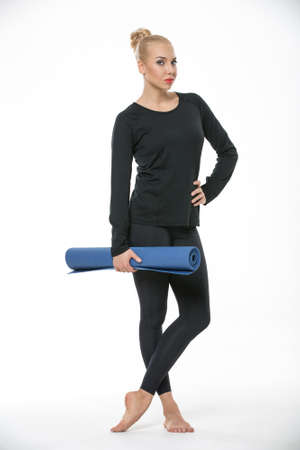 Pretty blonde girl in the sportswear stands barefoot on the white background in the studio. She wears black pants and black long sleeve t-shirt. She holds a blue gymnastic mat in the right hand. Her left hand is on the waist. Her legs are crossed. She looの写真素材