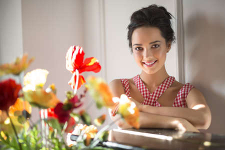 Smiling girl looks into the camera with crossed arms on the table. She is in the interior with light walls on background. There are flowers in front of her. Horizontal.の写真素材