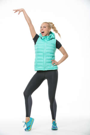 Sportive laughing blonde girl in the sportswear stands on the white background in the studio. She wears cyan-yellow sneakers, black-gray pants, black t-shirt and mint vest. She holds her left hand on her waist, right hand lifted up, right leg is on the toの写真素材