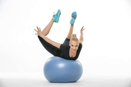 Gymnast girl in the sportswear lies on a blue fitball on the white background in the studio. She wears cyan-yellow sneakers, black pants and black t-shirt. She holds her stretched legs and arms in the air. She looks into the camera with a smile. Horizontaの写真素材