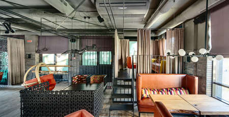 Restaurant in a loft style. On the right there is a wooden table with a brown sofa with motley pillow and white lamps over them, window with curtains. Behind them there is a zone on the metal elevation with stairs which framed with curtains. Inside the zoの写真素材
