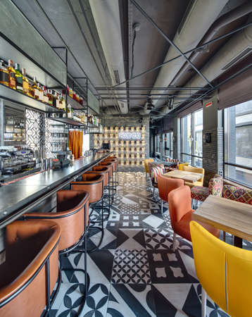Bar in a mexican restaurant in a loft style. On the left there is a black bar rack with brown chairs, shelves with dishes and equipment. On the right there are wooden tables with multi-colored chairs, windows with curtains. At the end there is a brick walの写真素材