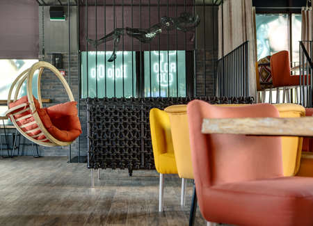 Interior in a mexican restaurant. There is a wooden table, multi-colored chairs, a black wicker sofa and a hanging wooden rocking chair with a red pillow. On the back wall there are windows with prints and curtains. On the right there is a zone framed witの写真素材