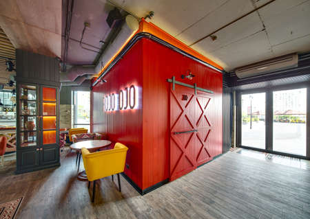 Hall in a loft style in a mexican restaurant. There is a red colored zone with a glowing inscription and an entrance to a restroom. On the left there are wooden tables with multi-colored chairs, a windows with curtains, a cupboard with glass sections. On の写真素材
