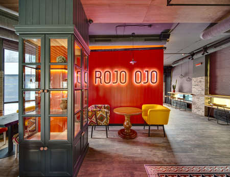 Interior in a loft style in a mexican restaurant. On the left there is a window with curtains, a cupboard with glass doors, wooden tables with multi-colored chairs. In the centre there is a red wall with glowing inscription, rounded wooden table with two の写真素材