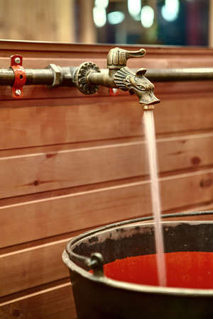 Metal faucet in the form of a fairy animal on the background of the wooden wall and mirror. Water flows from the faucet into a metal-red washbasin in the form of a bucket. Vertical.の写真素材