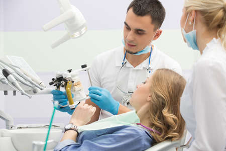 Dental cabinet with girl in blue shirt and patient bib on the patient chair. Near her sits a male dentist and a female assistant. They both wear white uniform with blue latex gloves and blue masks. Dentist also has binocular loupes. He holds an articulatoの写真素材