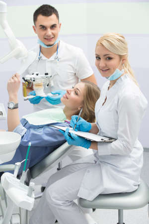 Attractive girl in blue shirt and patient bib on the patient chair in the dental cabinet. She holds a mirror in the left hand. Next to her sits a smiling male dentist and a smiling female assistant. They both wear white uniform with blue latex gloves and の写真素材