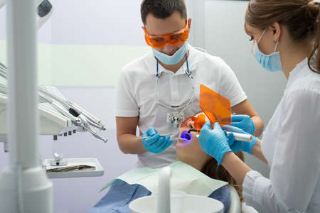 Female patient in blue shirt and patient bib on the patient chair in the dental cabinet. Next to her there is a male dentist and a female assistant. They both wear white uniform with blue latex gloves and blue masks. Patient and dentist wear UV protectiveの写真素材