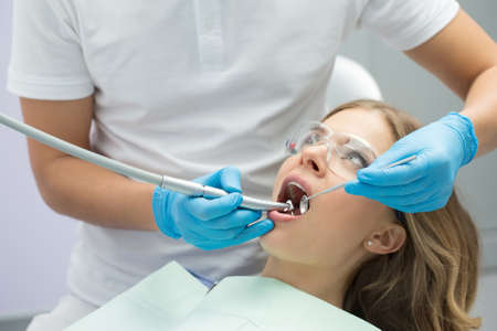 Brave girl with opened mouth in patient bib and protective eyewear. Next to her there is a dentist in a white uniform with blue latex gloves. He treats her teeth with a LED handpiece and a stem mouth mirror. Horizontal.の写真素材