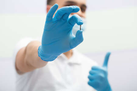 Dentist in a white uniform with blue latex gloves holds a dental implant in right hand. He shows like hand sign. Blurry background. Horizontal.の写真素材