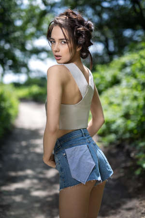 Cute girl stands on the nature background. She wears a silver top and denim shorts. She holds her...