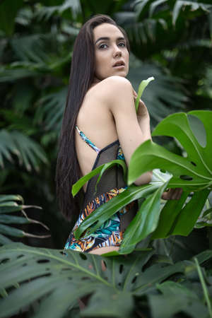 Wonderful girl stands sideways between big green plants. She wears a colorful swimsuit with pictures. She presses big leaf to her body with her hands. She looks up with parted lips. Vertical.の写真素材