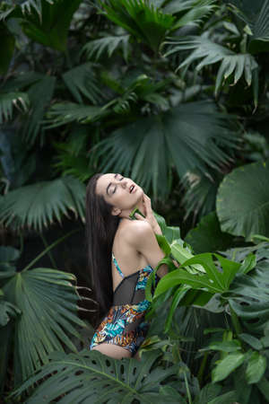 Sensuous girl stands sideways between big green plants. She wears a colorful swimsuit with pictures. She presses big leaf to her body with her hands. Her eyes are closed and mouth is open. Vertical.の写真素材