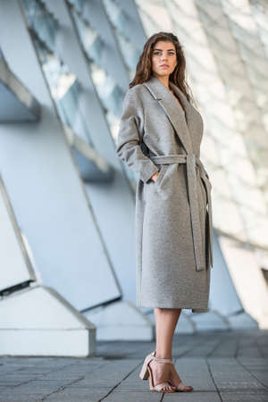 Amazing girl with beautiful hairstyle stands outdoors on the blurry urban background. She wears a...