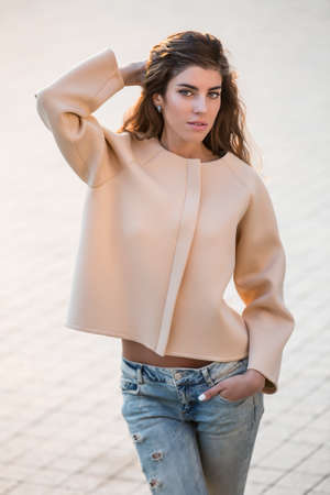 Cute girl with beautiful hairstyle stands outdoors on the blurry background. She wears a beige jacket and light ripped jeans. She holds her left hand in the pocket, right hand is behind the head. She looks into the camera with parted lips. Vertical.の写真素材