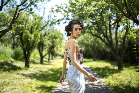 Stylish girl in a dress stands sideways on the nature background. Dress is with silver bottom and flowered top. She holds left hand along the body, right hand is bent at the elbow. She looks into the camera with parted lips. Horizontal.の写真素材