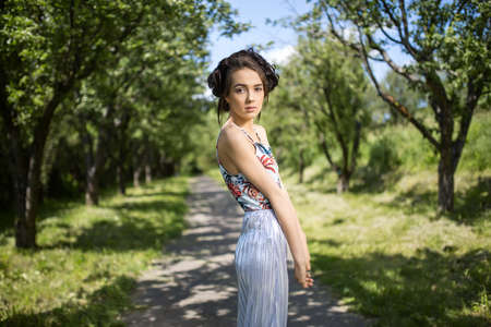 Young girl in a dress stands sideways on the nature background. Dress is with silver bottom and flowered top. She holds straightened hands in front of herself and looks into the camera. Horizontal.の写真素材