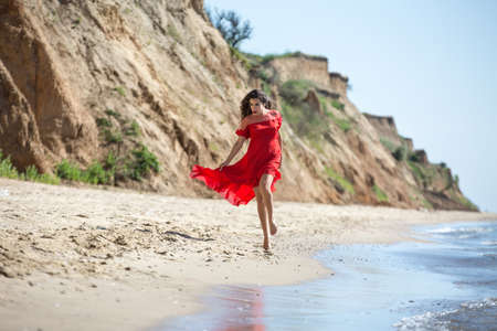 Attractive girl in a smart dress red goes along the seashore. Curly hair fluttering in the wind.の写真素材