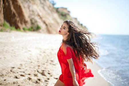 Pretty barefoot girl stands sideways on the sand on the cloudy sky background. She wears a white...
