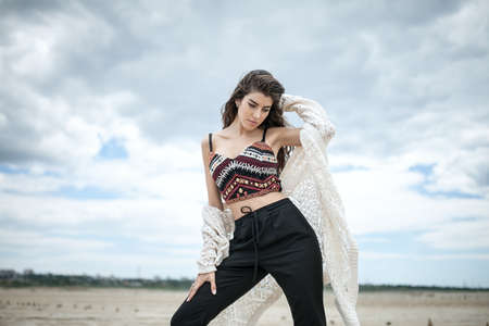 Thoughtful girl stands on the sand on the cloudy sky background. She wears black pants,...