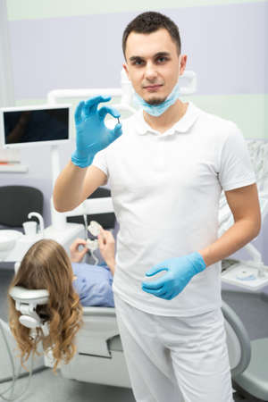 Handsome dentist in a white uniform with blue latex gloves and a blue mask holds an implant in the right hand. He stands on the background of dental equipment and a patient on the patient chair. Vertical.の写真素材