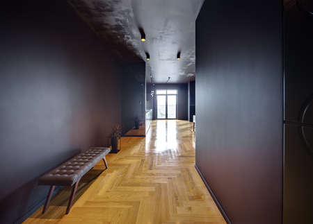 Hall in a modern style with dark walls and parquet on the floor. On the left there is a bench and a plant in the pot which reflected in the mirror wall of a kitchen. At the back there is a glass door.の写真素材