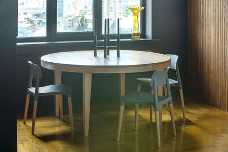 Wooden round table with three chairs on the background of the window with yellow vase and dark wall. Wooden wall is on the right. On the floor there is a parquet. On the table there is a candlestick.の写真素材