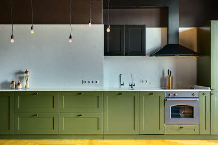 Kitchen in a modern style with a light tabletop with sink, cooker, oven, kitchen accessories. Under tabletop there are green drawers. Over tabletop there is kitchen hood, cupboard and glowing lamps.の写真素材