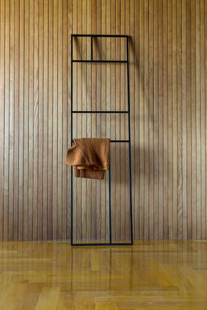 Black metal frame stand which leans on the wooden wall. Brown pullover hangs on the stand. On the floor there is a parquet. Indoors. Vertical.の写真素材