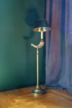 Design golden metal glowing lamp with turquoise shade on the background of the turquoise wall and curtain. Lamp drops shadow on the wall. On the floor there is a parquet. Indoors. Vertical.の写真素材