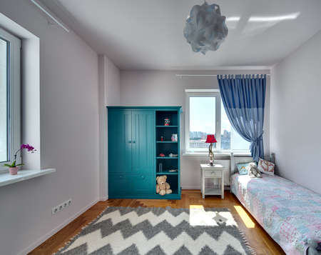 Room for kid with light walls and a parquet with carpet on the floor. There is a bed with colorful veil and pillows and a toy, turquoise bookcase with toys and books, nightstand with a lamp.の写真素材