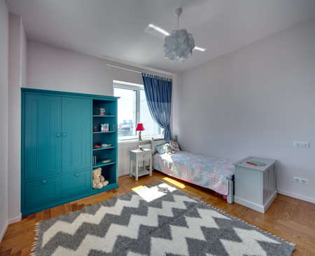 Kid's bedroom with light walls and a parquet with carpet on the floor. There is a bed with colorful veil and pillows and a toy, turquoise bookcase with toys and books, nightstand with a lamp, commode.の写真素材
