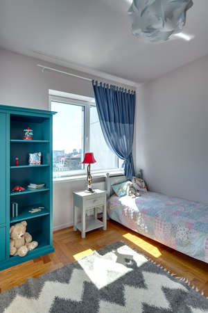 Children's bedroom with white walls and a parquet with carpet on the floor. There is a bed with colorful veil and pillows and a toy, turquoise bookcase with toys and books, nightstand with a lamp.の写真素材