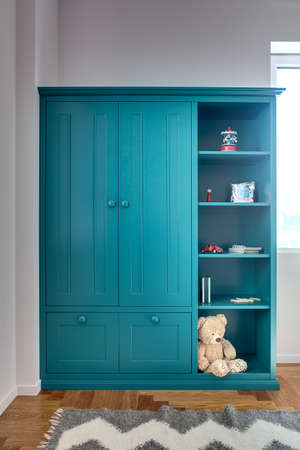 Kid's turquoise bookcase with toys, books, notebooks, pencils and a big teddy bear. On the floor there is a parquet with a gray carpet. Walls are white. Indoors. Vertical.の写真素材