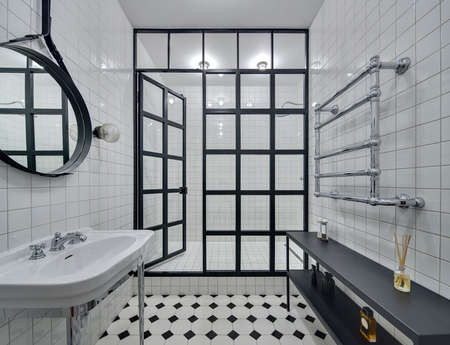 Bathroom with walls of white tiles. There is white washbasin, black mirror, shower with glass partition with open door and glowing lamps, black rack with accessories and chromed heated towel rail.の写真素材