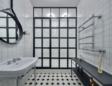 Bathroom with walls of white tiles. There is white washbasin, black mirror, shower with black partition of glass squares and glowing lamps, black rack with accessories and chromed heated towel rail.の写真素材