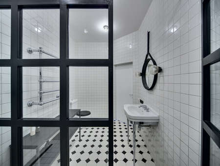 Modern bathroom with walls of white tiles. There is white washbasin, black mirror, lamps, glass partition, white-black toilet, black rack with accessories, chromed heated towel rail, white door.の写真素材