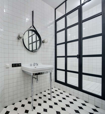 Design bathroom with walls of white tiles. There is white washbasin with chromed legs and faucet, black mirror with two lamps, shower with black partition of glass squares and glowing lamp.の写真素材
