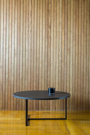 Black wooden round table with metal legs on the background of the wooden wall. There is a dark glass box on the table. On the floor there is a parquet. Indoors. Horizontal.の写真素材