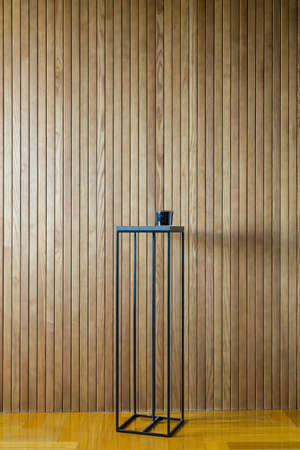 Dark metal stand on the background of the wooden wall. There is a dark glass box on the stand. On the floor there is a parquet. Indoors. Vertical.の写真素材