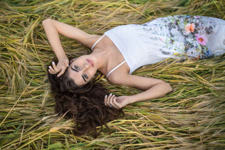 Sexy girl lies on her back on the rye and looks into the camera with a smile. She wears a light dress with prints of flowers. Her hands are on the hair. Outdoors. Horizontal.の写真素材