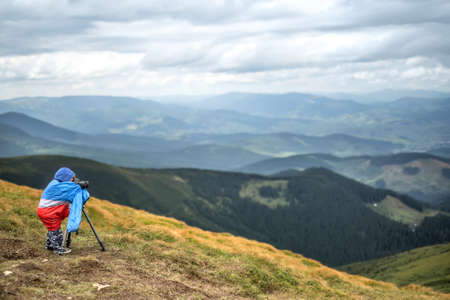 Cute kid in multi-colored big size windcheater is making a photo on the background of mountains and the cloudy sky. He is looking into the viewfinder of the camera which is on the tripod. Horizontal.の写真素材