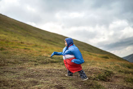 Funny kid in multi-colored big size windcheater walks on the mountain on the cloudy sky background. Shoot from the side. Horizontal.の写真素材