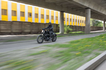 Guy in black helmet hurtling on the black motorbike on the road under the overpass. He wears a black leather jacket, black jeans, black shoes, black gloves and sunglasses. Outdoors. Horizontal.の写真素材