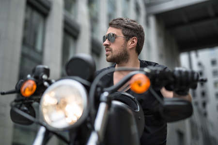 Charismatic man sits on the black motorbike on the skyscraper background. Headlamp switched on. He wears a black T-shirt, black gloves and sunglasses. He looks to the right with parted lips. He holds hands on the rudder. Black helmet lies on the motorbikeの写真素材