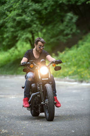 Stylish man with a beard rides on the black motorbike on the green tree background. Headlamp...