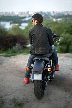 Rider sits on the black motorbike on the background of the river and the city. He looks to the left. He wears a blue ripped jeans, red sneakers, a black leather jacket and sunglasses. Photographed from the back. Outdoors. Vertical.の写真素材