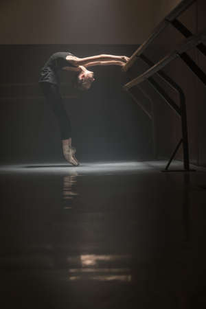 Slim ballerina standing in a position next to ballet barre. The light falls from above on her face, arms and body. Low key photo.の写真素材