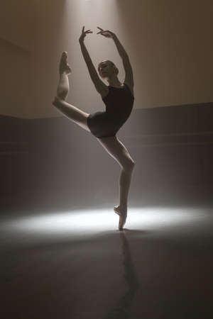 Pretty ballerina posing during her training in the dark dance hall. She standing on one leg with raised hands and other leg. Light falls down from above behind her. Shoot in a low key.の写真素材